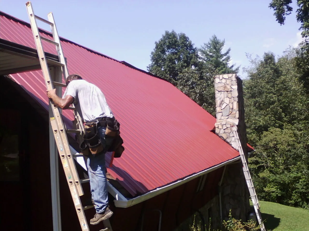 Roofer on a ladder beside a red metal roof and stone chimney — Soffit & Fascia Repair by Roofing Brothers in Clitherall